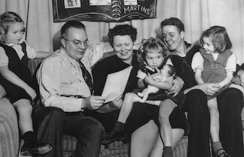 Una familia de seis personas, incluyendo dos adultos, un joven y tres niñas, sentada en un sofá, sonriendo. El padre lee un papel, una niña sostiene una muñeca