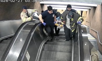 Vista desde arriba de personal de emergencia, con uniformes de bombero y paramédico, bajando una camilla con una persona por una escalera mecánica metálica gris