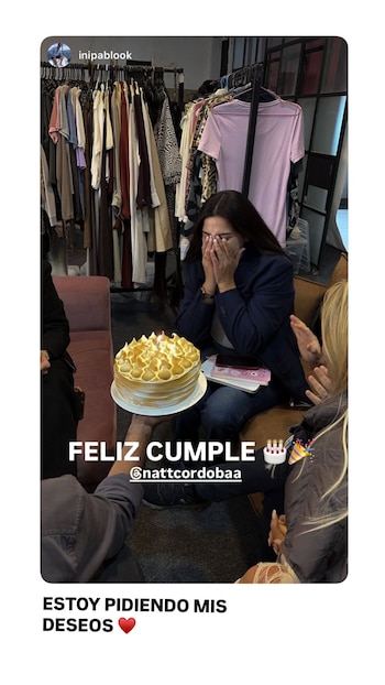 Una mujer con cabello oscuro y chaqueta azul se cubre el rostro con las manos, mientras le presentan una tarta de cumpleaños con merengue y una vela encendida