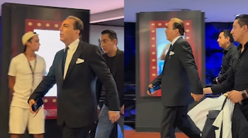 Un hombre con traje oscuro y corbata negra camina acompañado por varias personas. Un joven usa gorra y camiseta blanca. Hay pantallas iluminadas al fondo.