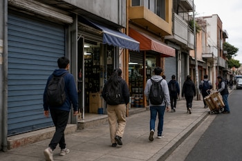 Jóvenes con mochilas caminan de espaldas por una acera junto a locales comerciales con persianas metálicas.