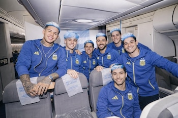 Grupo de siete hombres jóvenes con chaquetas azul y amarillo de la Selección Argentina, sonriendo en el interior de un avión, con el cabello teñido de azul y blanco.