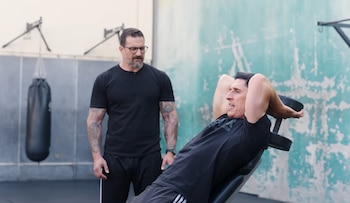 Jeff Cavaliere está acostado en un banco inclinado, levantando una mancuerna sobre su cabeza, mientras un hombre barbudo lo mira de cerca en un gimnasio