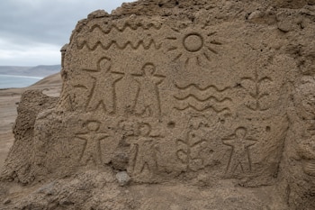 Grabados en un muro de barro con figuras humanas esquemáticas, líneas onduladas y un sol sobre un paisaje árido de fondo.