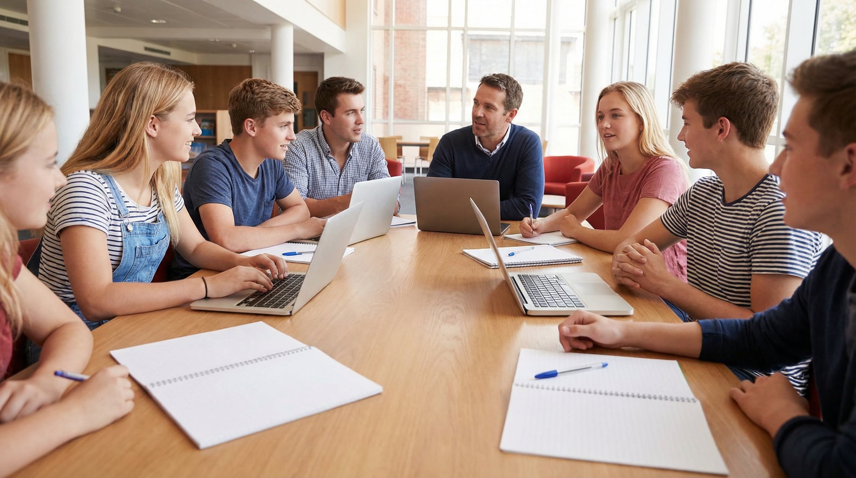 Un grupo diverso de estudiantes adolescentes y adultos, junto a un profesor, participan en un animado debate en una mesa equipada con laptops y cuadernos. (Imagen Ilustrativa Infobae)