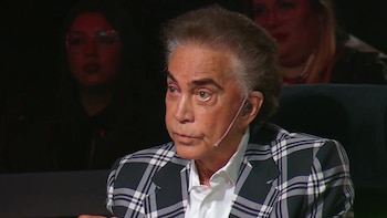 José Luis Rodríguez viste un saco a cuadros y lleva un micrófono de diadema mientras mira hacia la izquierda en un estudio de televisión