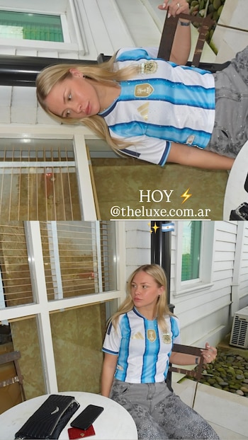 Mujer rubia sentada viste camiseta de la selección argentina y pantalón gris, frente a una mesa blanca con cartera, teléfono y tarjetero. Edificio blanco
