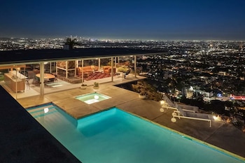 Casa moderna de cristal con piscina iluminada y terraza sobre una colina frente a la vista nocturna de una ciudad