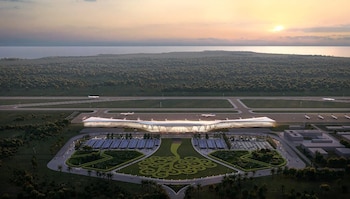 Vista aérea de una terminal aeroportuaria con pistas de aterrizaje, aviones estacionados, zonas verdes y el mar al fondo durante el atardecer