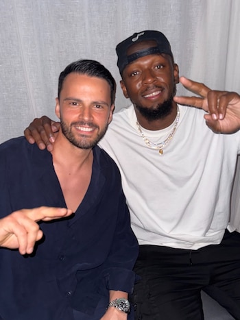 Dos hombres sonrientes posan juntos, uno con camisa oscura y el otro con camiseta blanca, gorra y collares, con cortinas claras de fondo
