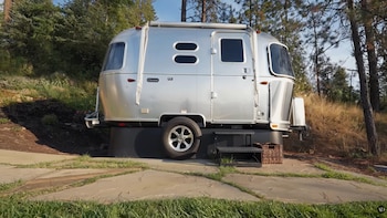 Remolque Airstream plateado brillante sobre una plataforma oscura, con una rueda visible, escalones portátiles y una cesta de mimbre, rodeado de árboles y arbustos