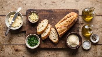 Vista superior de una baguette rebanada en una tabla de madera junto a cuencos con mantequilla, ajo picado, perejil, queso y aceite de oliva.