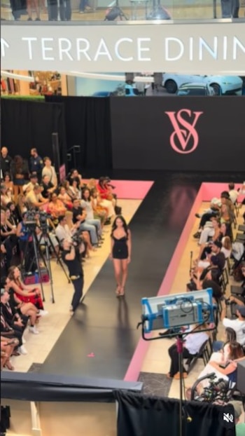 Mujer de cabello oscuro en vestido negro camina por pasarela oscura con bordes rosados, frente al logo de VS en pantalla y espectadores