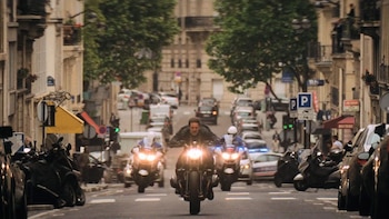Un hombre en una motocicleta oscura acelera por una calle urbana rodeada de edificios, seguido por dos motocicletas con luces brillantes y varios coches aparcados