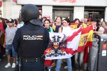 Un agente de policía de espaldas con casco y chaleco táctico observa a un grupo de personas que sostienen banderas de España