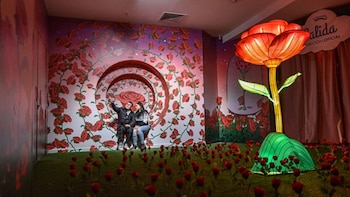 Dos personas, hombre y mujer, se toman una selfi sentados en un banco con fondo de rosas rojas, césped artificial y una flor gigante iluminada