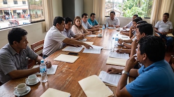Un grupo de catorce personas sentadas alrededor de una mesa de madera. Hay documentos, carpetas, botellas de agua, tazas de café y teléfonos móviles sobre la mesa. Ventanas con vista al exterior
