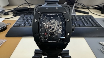 Reloj de pulsera negro con mecanismo visible sobre un soporte, ubicado frente a un teclado de computadora y herramientas de trabajo.