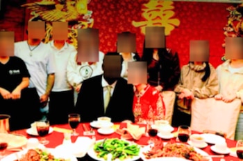 Grupo de personas en una mesa con mantel rojo, copas de vino y platos de comida. Un fondo rojo tiene símbolos chinos dorados