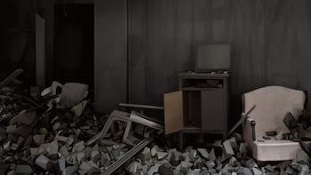 Un cuarto oscuro con montones de escombros grises, un televisor antiguo sobre un mueble, un sillón claro, y objetos fragmentados en el suelo