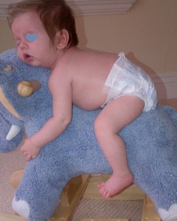Bebé dormido con pañal sobre un balancín de peluche azul con forma de elefante. Un corazón azul cubre la cara del bebé