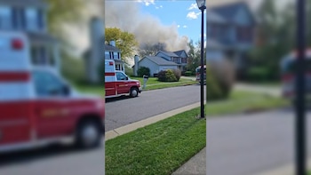 Una ambulancia roja y blanca de Akron Fire y un camión de bomberos en una calle residencial; densas nubes de humo se elevan sobre las casas al fondo