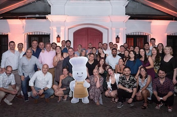 Grupo numeroso de hombres y mujeres de diversas edades posa sonriendo al aire libre con un recorte de cartón de un chef con delantal y nombre Hudson
