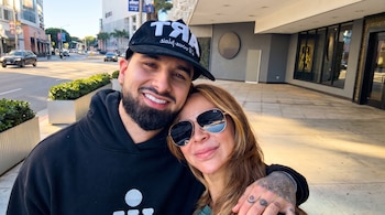 Hombre con barba y gorra negra abrazando a una mujer con pelo castaño y gafas de sol, posando en la acera de una calle urbana con edificios.