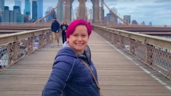 Mujer con cabello rosa y chaqueta azul oscuro sonríe en un puente con pasarela de madera y estructura metálica, con edificios de una ciudad de fondo