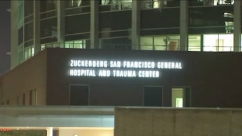 Fachada nocturna de un edificio moderno de hospital con ventanales y un gran letrero luminoso que dice: Zuckerberg San Francisco General Hospital and Trauma Center