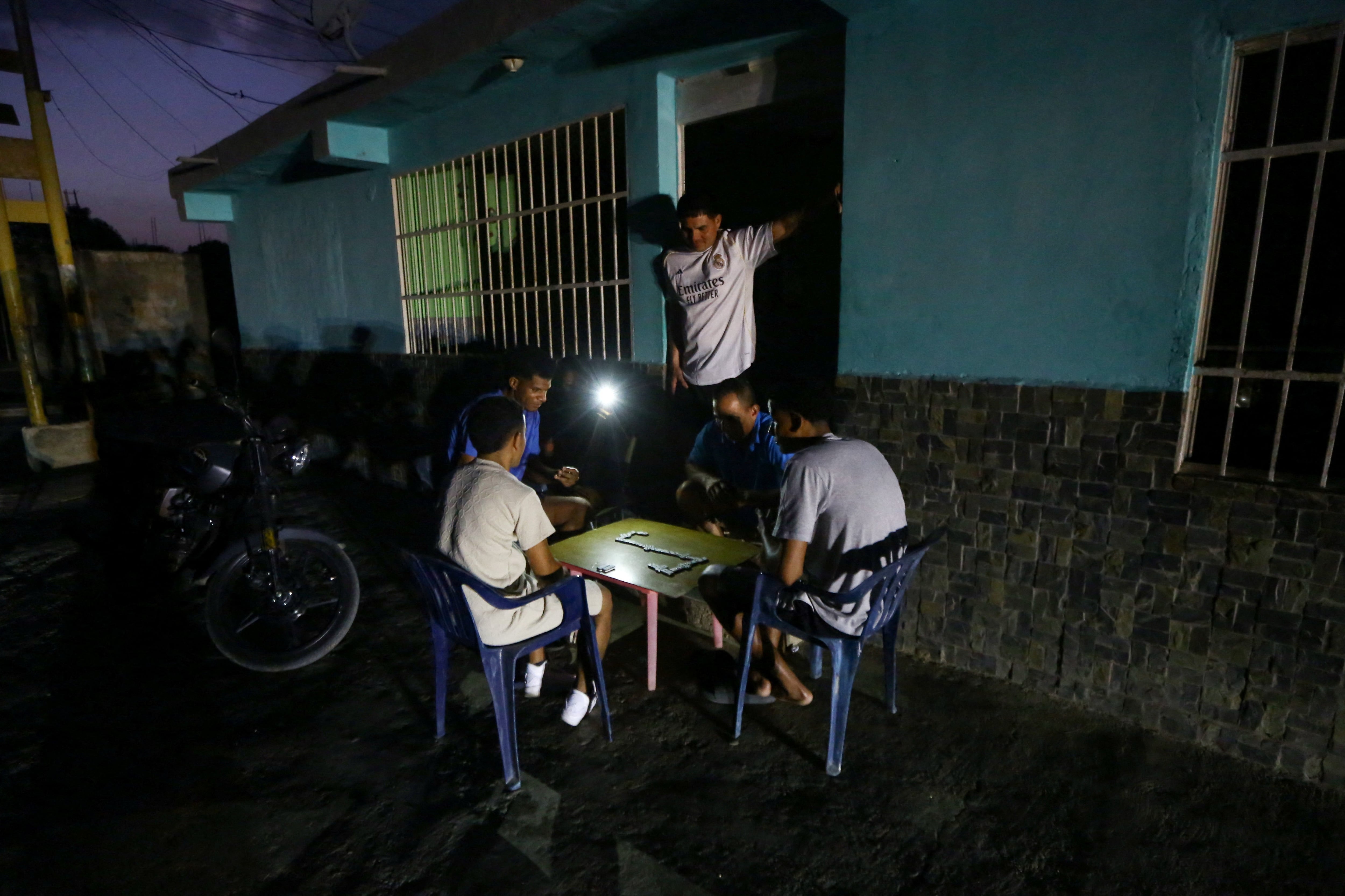 Unas personas juegan al dominó a la luz de un teléfono móvil durante un apagón, mientras los venezolanos enfrentan cortes de electricidad cada vez más frecuentes debido al deterioro de la red eléctrica del país (REUTERS/Juan Carlos Hernandez/Archivo)