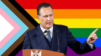 Hombre en traje azul habla en un podio. Detrás, la bandera Progress Pride con franjas arcoíris, rosa, azul claro, blanco, marrón y negro.