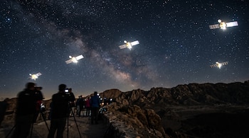 Siluetas de personas con cámaras en un entorno rocoso bajo un cielo estrellado con cuatro satélites artificiales iluminados.