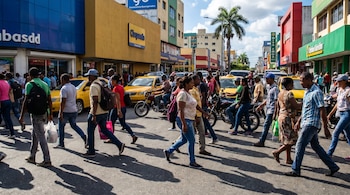 Multitud de peatones cruza una avenida en República Dominicana, con tráfico vehicular, edificios coloridos y una palmera bajo un cielo azul caribeño.