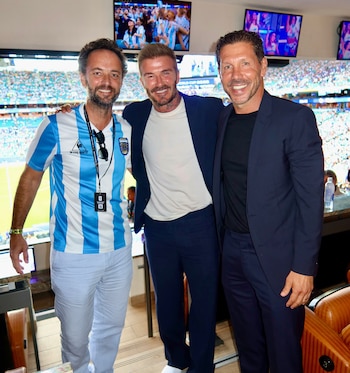 Marco Materazzi, David Beckham y Diego Simeone posan sonrientes en el palco de un estadio con el campo de fútbol y el público de fondo. Hay pantallas en la parte superior