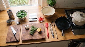 Utensilios de cocina, tablas de cortar, un cuchillo, sartén, olla, báscula e ingredientes organizados sobre una encimera de madera.