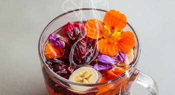 Taza de tisana caliente con flores comestibles naranjas, violetas y rodajas de banana vista desde arriba
