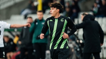 Un jugador de fútbol con camiseta negra y verde de manga larga camina en la cancha con personas fuera de foco al fondo.