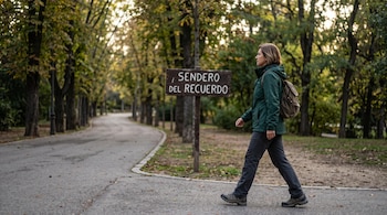 Una mujer de perfil con abrigo verde y mochila camina por un sendero junto a un cartel de madera grabado con texto.