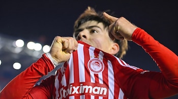 Un jugador de fútbol con la camiseta de rayas rojas y blancas del Olympiacos sostiene su uniforme cerca del rostro y apunta hacia arriba.