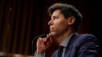 Primer plano de perfil de Sam Altman, vestido con un traje azul y corbata estampada, mirando pensativo hacia la derecha, con un micrófono delante