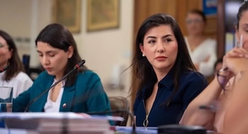 Mujeres en una conferencia académica, sentadas en una mesa con micrófonos. Una mujer de cabello oscuro y blusa azul mira a la derecha; otra de blazer turquesa mira hacia abajo