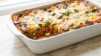 Bandeja blanca rectangular con lasagna vegetariana, capas de vegetales salteados, salsa de tomate y queso gratinado, decorada con hojas de cilantro.