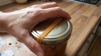 Una mano sujeta la tapa metálica de un frasco de vidrio con una banda elástica de goma en el borde, sobre una encimera de madera.