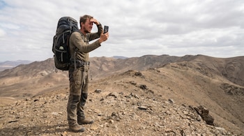 Un hombre con mochila grande y ropa de senderismo se para en la cima de una montaña rocosa, mirando su teléfono con expresión de frustración bajo un cielo nublado.