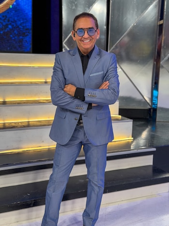 Hombre con traje azul, camisa negra y gafas de pie con los brazos cruzados frente a unas gradas iluminadas en un estudio de televisión