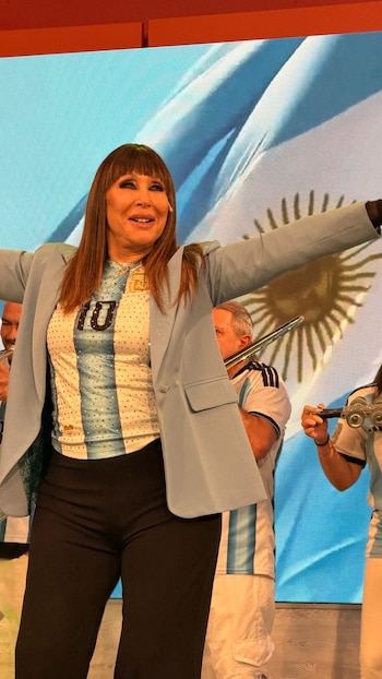 Moria Casán, con camiseta celeste y blanca con el número diez y saco celeste, frente a una pantalla con la bandera argentina. Hay otras personas detrás
