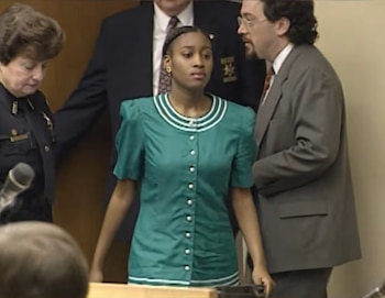 Una mujer con vestido verde camina en una sala judicial acompañada por un hombre con traje y una agente policial