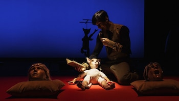 Un hombre de rodillas con micrófono y libro junto a una marioneta con orejas de gato. Dos cabezas de maniquí en almohadas. Fondo azul con silueta de sombra