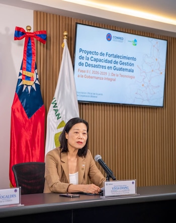 Mujer de cabello oscuro con traje beige sentada en mesa negra con micrófono, frente a dos banderas y una pantalla con presentación de texto en español
